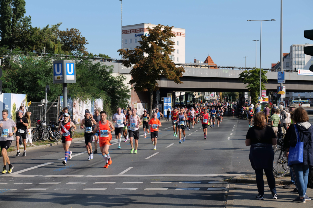 Gruppe von Läufern bei einem Marathon auf einer von Bäumen gesäumten Straße mit Fahrrädern, Schildern, einem Zaun, einer Brücke, Gebäuden und einem klaren blauen Himmel.