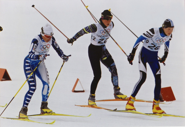 Drei Skifahrer in buntfarbenen Anzügen und Helmen sausen eine glitzernde schneebedeckte Piste hinunter, jeder hält Ski-Stöcke.