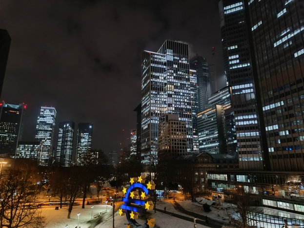 Europäische Zentralbank-Hauptquartier in Frankfurt, Deutschland bei Nacht mit beleuchteten Gebäuden, Bäumen, Straßenlaternen, Lichtern und Fußgängern unter bewölktem Himmel.