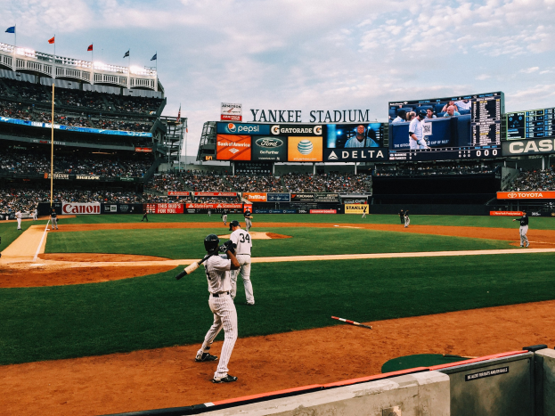 Baseballspiel im Yankee Stadium mit Spielern auf dem Feld, Zuschauern in den Rängen und Stadionmerkmalen wie einem Zaun, Fahnen, Anzeigetafeln, einem Bildschirm und Deckenleuchten unter einem bewölkten Himmel.