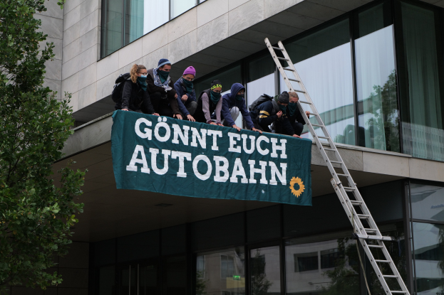 Gruppe von Menschen auf einem Gebäudedach mit einer Fahne, mit einem Baum links und Glasfenstern im Hintergrund, die scheinbar für die deutsche Regierung protestieren.