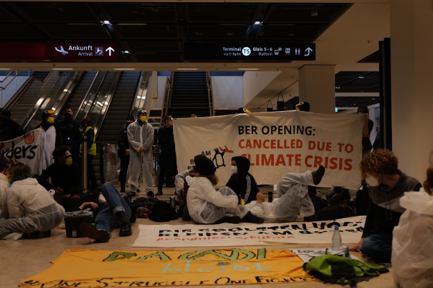Eine Gruppe von Menschen sitzt auf dem Boden eines Flughafens und hält ein Banner mit der Aufschrift "Berlin aufgrund des Klimawandels abgesagt", umgeben von verstreuten Taschen, Flaschen und anderen Gegenständen sowie Rolltreppen, Texttafeln und Deckenleuchten im Hintergrund.