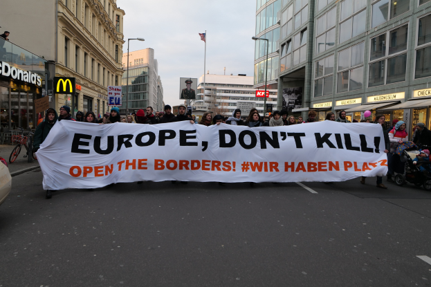 Eine Gruppe von Menschen marschiert mit einem Banner, auf dem "Europa, töte nicht, öffne die Grenzen" steht, die Straße entlang, mit einem geparkten Auto auf der linken Seite, Gebäuden und einer Flagge im Hintergrund unter einem klaren blauen Himmel.