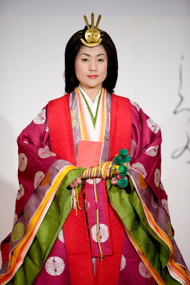Eine Frau in einem traditionellen japanischen Kimono, das ein leuchtend rotes und grünes Kleid mit aufwändigen Mustern zeigt, steht selbstbewusst auf einem Laufsteg, ihr Haar ordentlich zu einem Knoten gebunden.