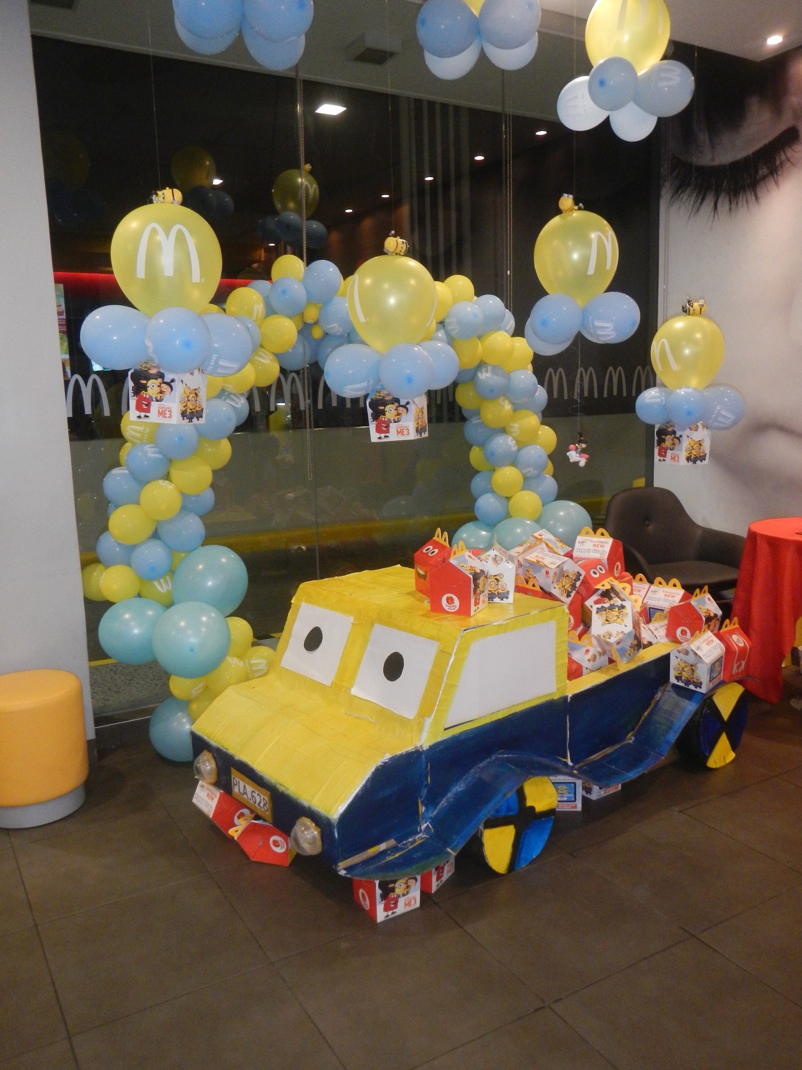 Eine festliche McDonald's-Geburtstagsparty-Szene mit einem gelben und blauen Lastwagen, der mit Ballons und anderen Dekorationen geschmückt ist, einem Tisch mit einer roten Decke und Gegenständen darauf, Hockern und einer Glaswand im Hintergrund, unter Deckenlampen.