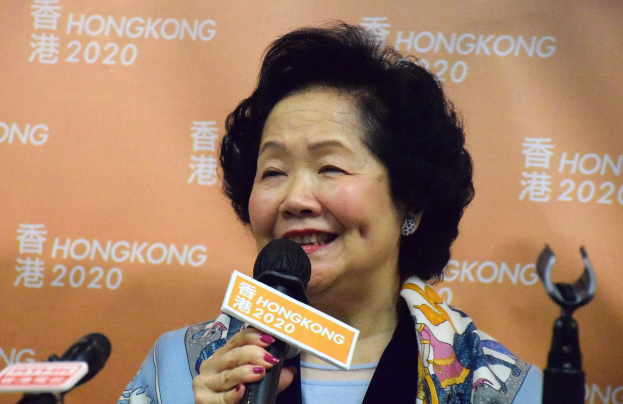 Hongkonger Chief Executive Carrie Lam bei einer Pressekonferenz, mit einem Mikrofon in der Hand und mehreren Mikrofonen vor ihr, sowie einem orangefarbenen Banner mit Text im Hintergrund.