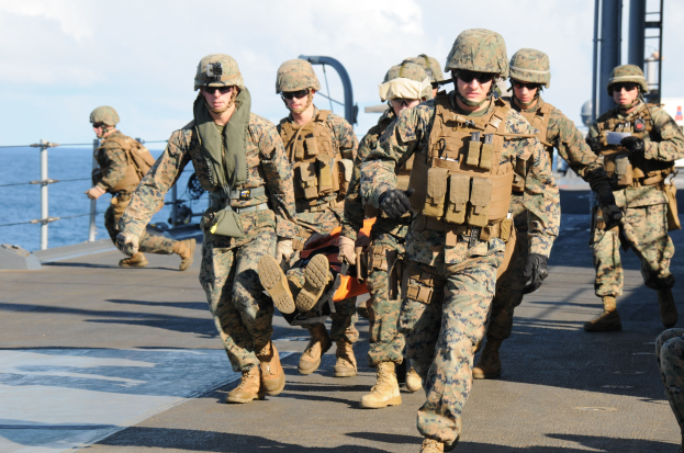 Marines in voller Ausrüstung tragen eine Person auf einer Trage über das Deck eines Schiffs mit Wasser und Himmel im Hintergrund.