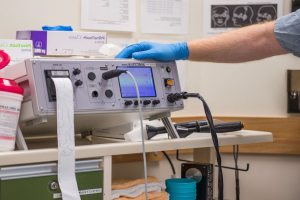 Eine Person in blauen Handschuhen arbeitet an einem medizinischen Gerät auf einem Tisch, der mit verschiedenen Gegenständen überhäufte ist, mit Papieren an der Wand im Hintergrund, was eine Krankenhaus\-Atmosphäre vermuten lässt.