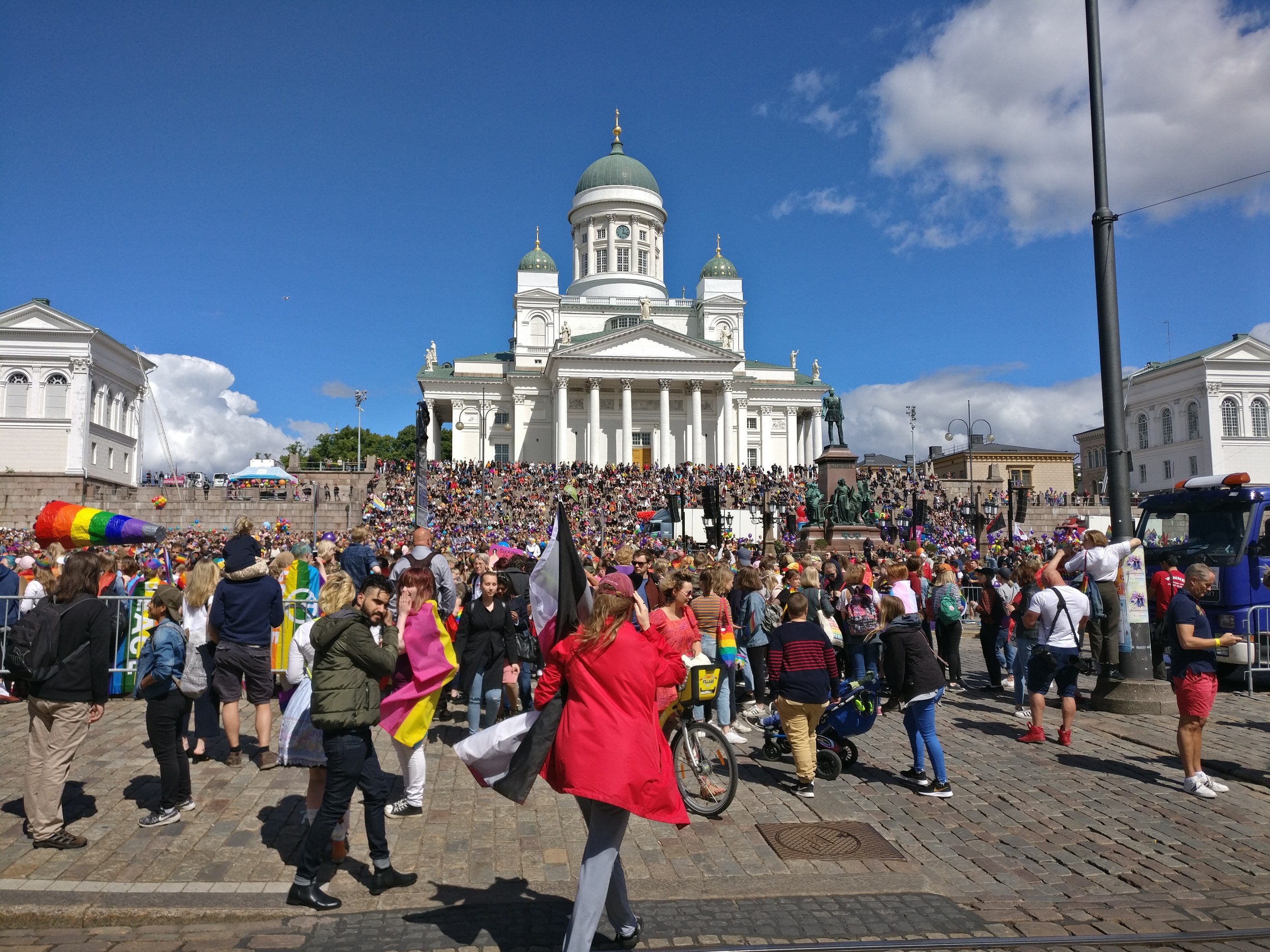 A large crowd walks down a street past a building with windows, pillars, and arches, some holding flags or pushing strollers, with parked vehicles, light poles, trees, and a cloudy sky in the background.