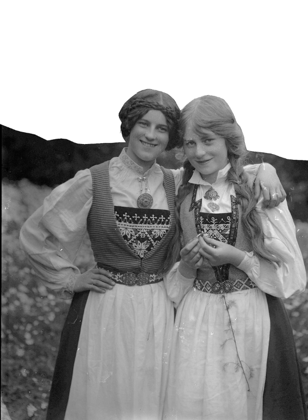 Zwei junge Frauen in traditionellen bayerischen Trachtenkleidern, die weiß und schwarz mit aufwendiger Stickerei und Perlen verziert sind, posieren vor der Kamera und lächeln, vor einem Hintergrund aus üppigen grünen Pflanzen und einem hellen weißen Himmel.