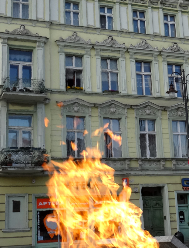 Ein großes Feuer brennt vor einem Gebäude mit Fenstern, Türen, einem Laternenmast und einer Tafel mit Schrift darauf.