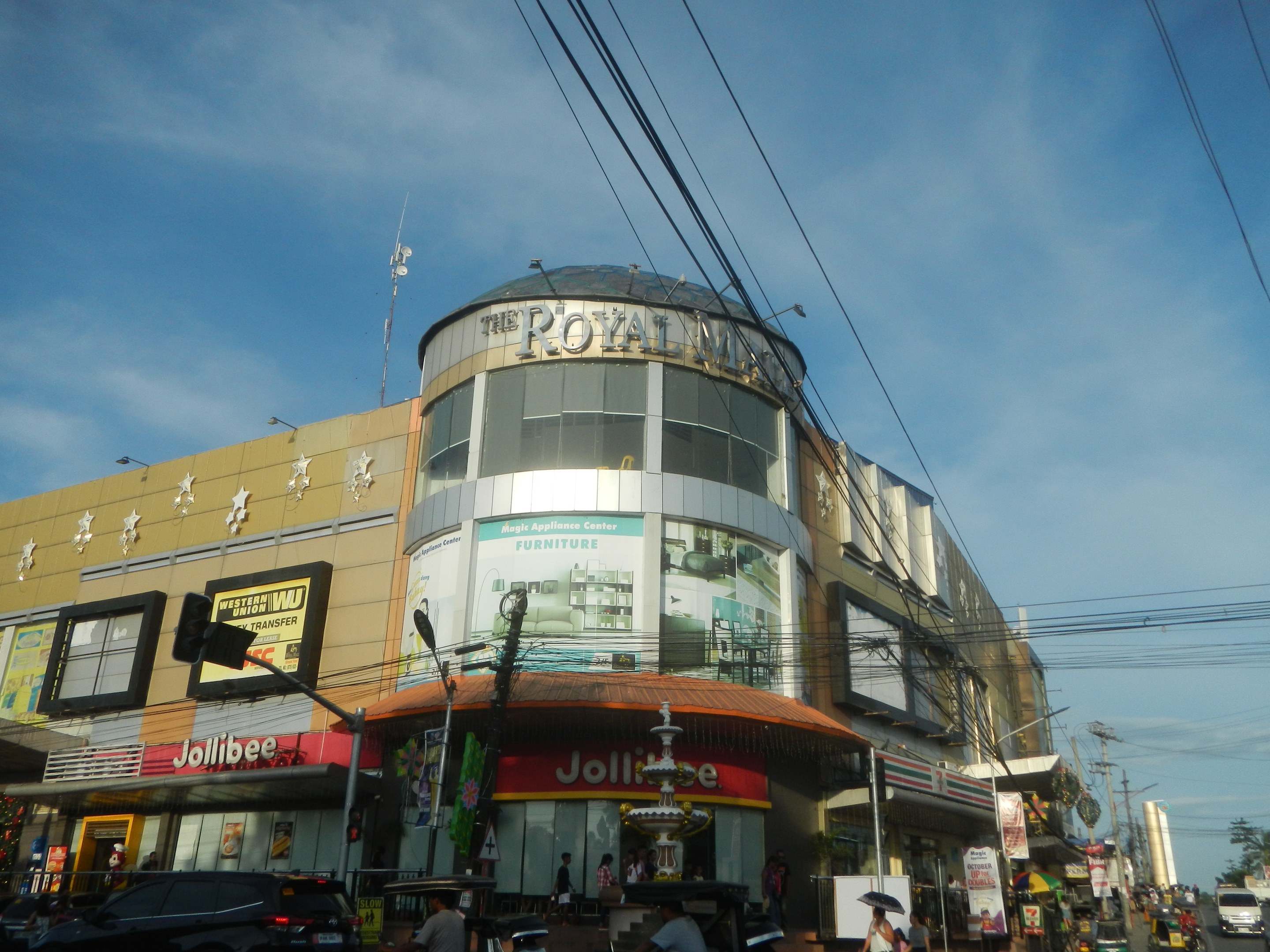 Eine belebte Stadtstraße mit fahrenden Fahrzeugen, Fußgängern auf dem Gehweg und dem Royal Mall-Einkaufszentrum im Hintergrund, umgeben von Bäumen, Strommasten und Drähten unter einem Himmel.