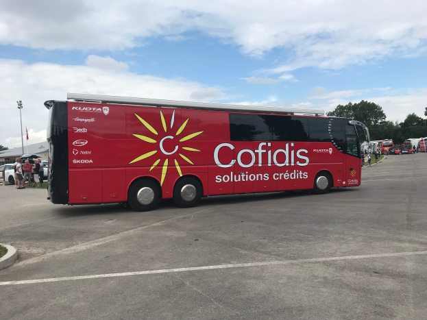 Ein roter Bus ist in einem Parkplatz neben einem Gebäude geparkt, mit Menschen, Fahrzeugen, einem Straßenschild, einem Schild mit der Aufschrift "Cofidis Solutions Credit", Bäumen und einem bewölkten Himmel im Hintergrund.