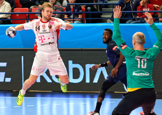 Eine Gruppe von Männern spielt Handball auf einem Feld mit einem Ball in der Mitte, während Zuschauer von Stühlen und Geländern in einem Stadion zusehen, mit einer Tafel im Hintergrund, auf der "Futsal-Weltmeisterschaft 2019 - Bayern München gegen Paris Saint-Germain" steht.
