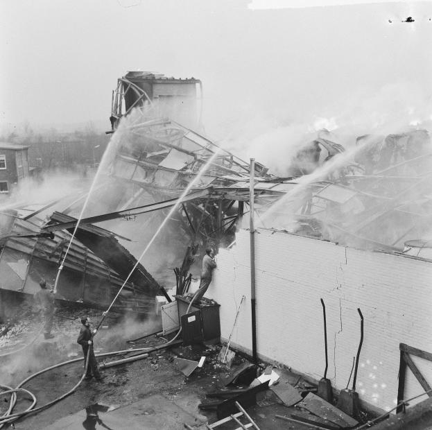 Schwarzes und weißes Foto von Feuerwehrleuten auf einem Dach, die ein Feuer löschen, während Zuschauer unten Ausrüstung halten.