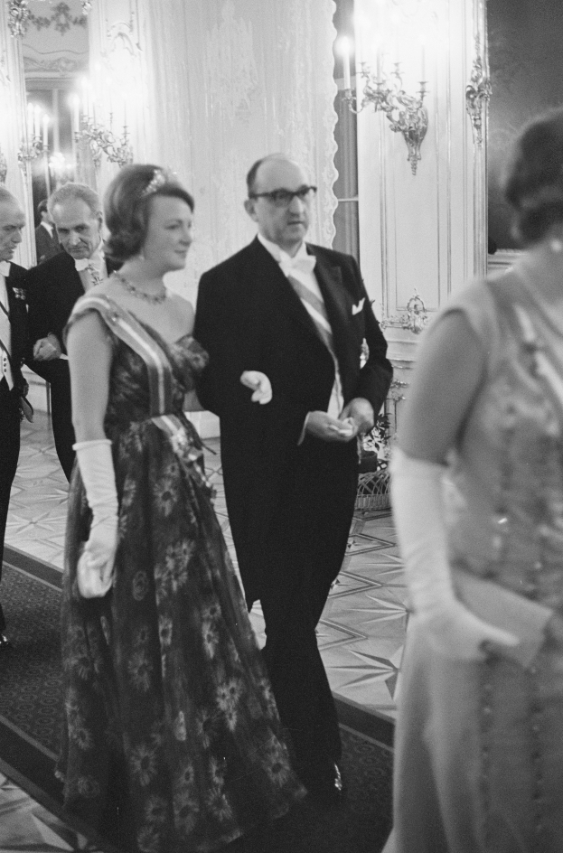 Ein Mann in einem schwarzen Blazer, weißem Hemd und schwarzen Hosen sowie eine Frau in einem eleganten Kleid gehen einen Flur entlang, umgeben von einer Gruppe von Menschen, die auf dem Boden stehen, während einige an der Wand montierte Lampen eine gemütliche Atmosphäre schaffen.