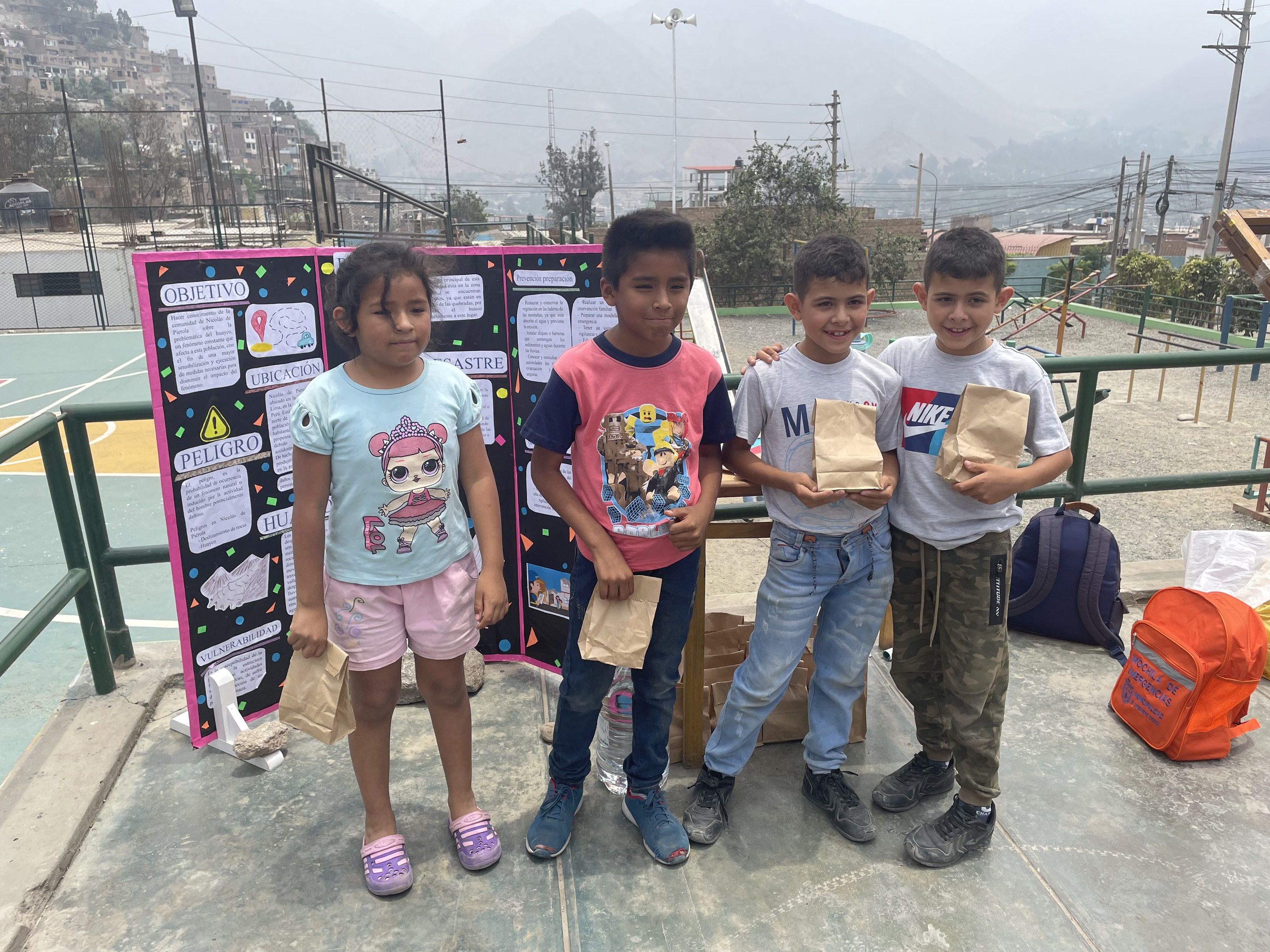 Gruppe von Kindern auf einem Basketballfeld mit Papiertüten in den H├Ąnden, mit Tüten und Gegenst├Ąnden rechts und einer Tafel mit Text links, vor dem Hintergrund von Z├Ąunen, Pfosten, Stra├čenlaternen, B├Ąumen, Geb├Ąuden, H├Ąngen und Himmel.