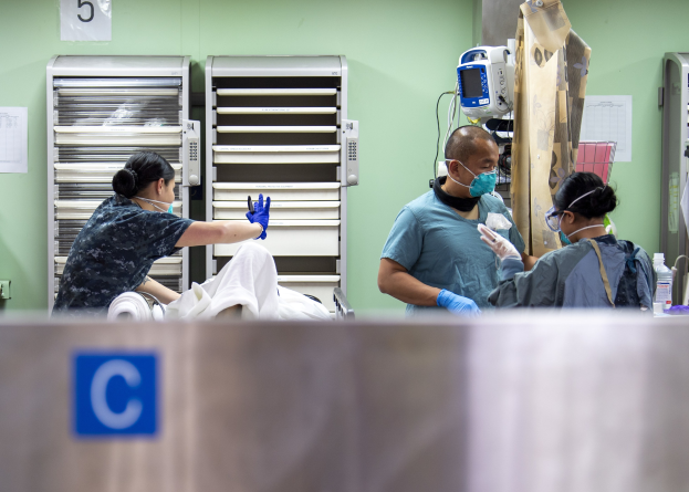Medical professionals in scrubs and masks attending to a patient on a draped hospital bed, with medical supplies on a nearby table and equipment in the background.