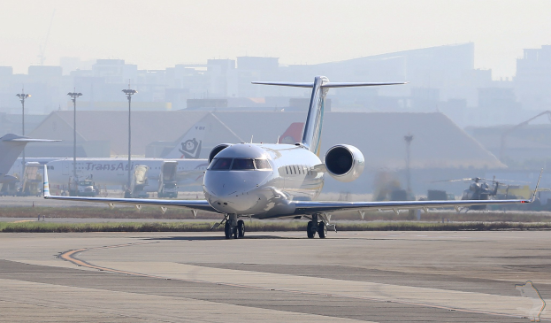 Private jet on an airport runway with surrounding infrastructure and clear blue sky.