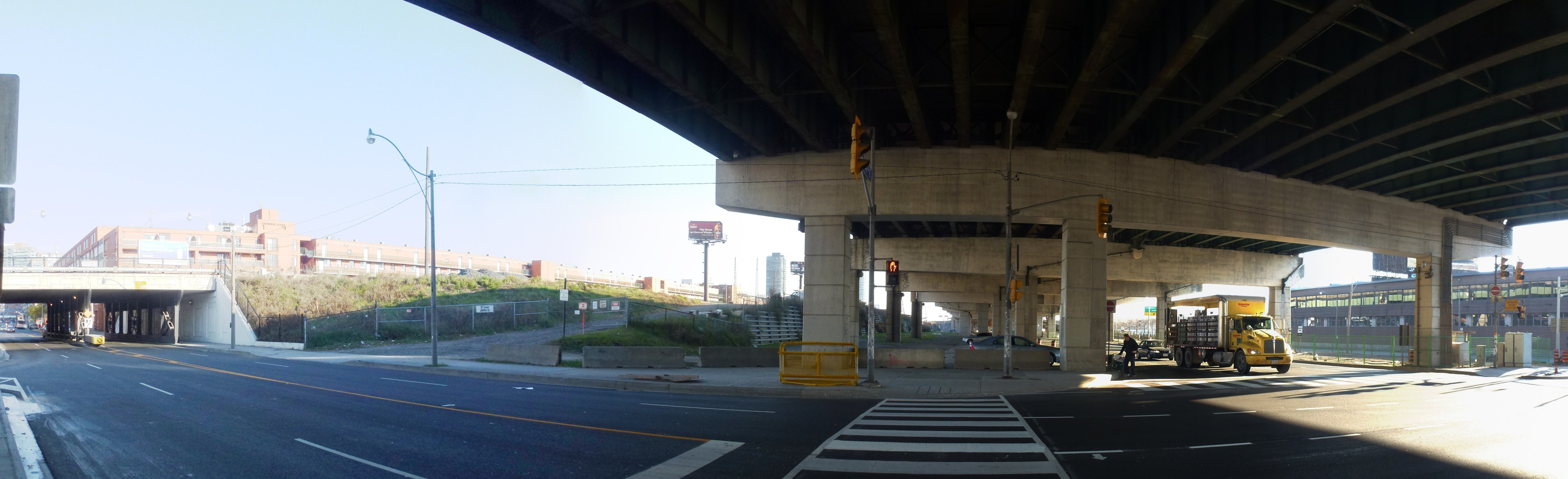 A truck driving under a bridge on a city street with buildings, street furniture, vehicles, and greenery under a clear sky.