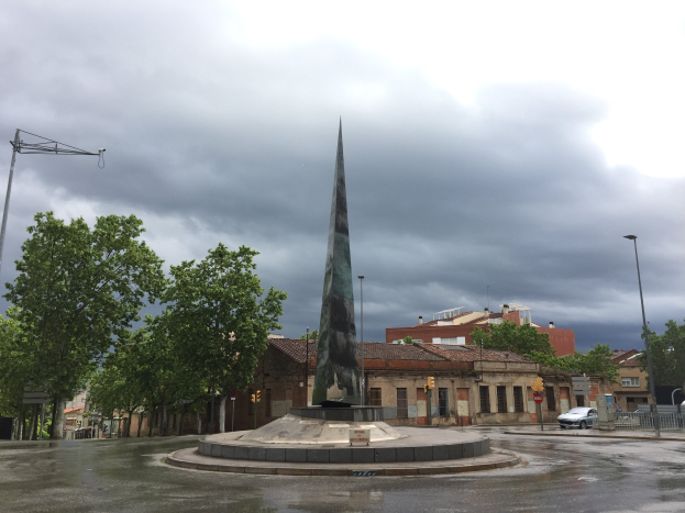 Ein Denkmal steht auf einem Stadtplatz umgeben von Gebäuden, Bäumen, Straßeninfrastruktur, Fahrzeugen und einem bewölkten Himmel.