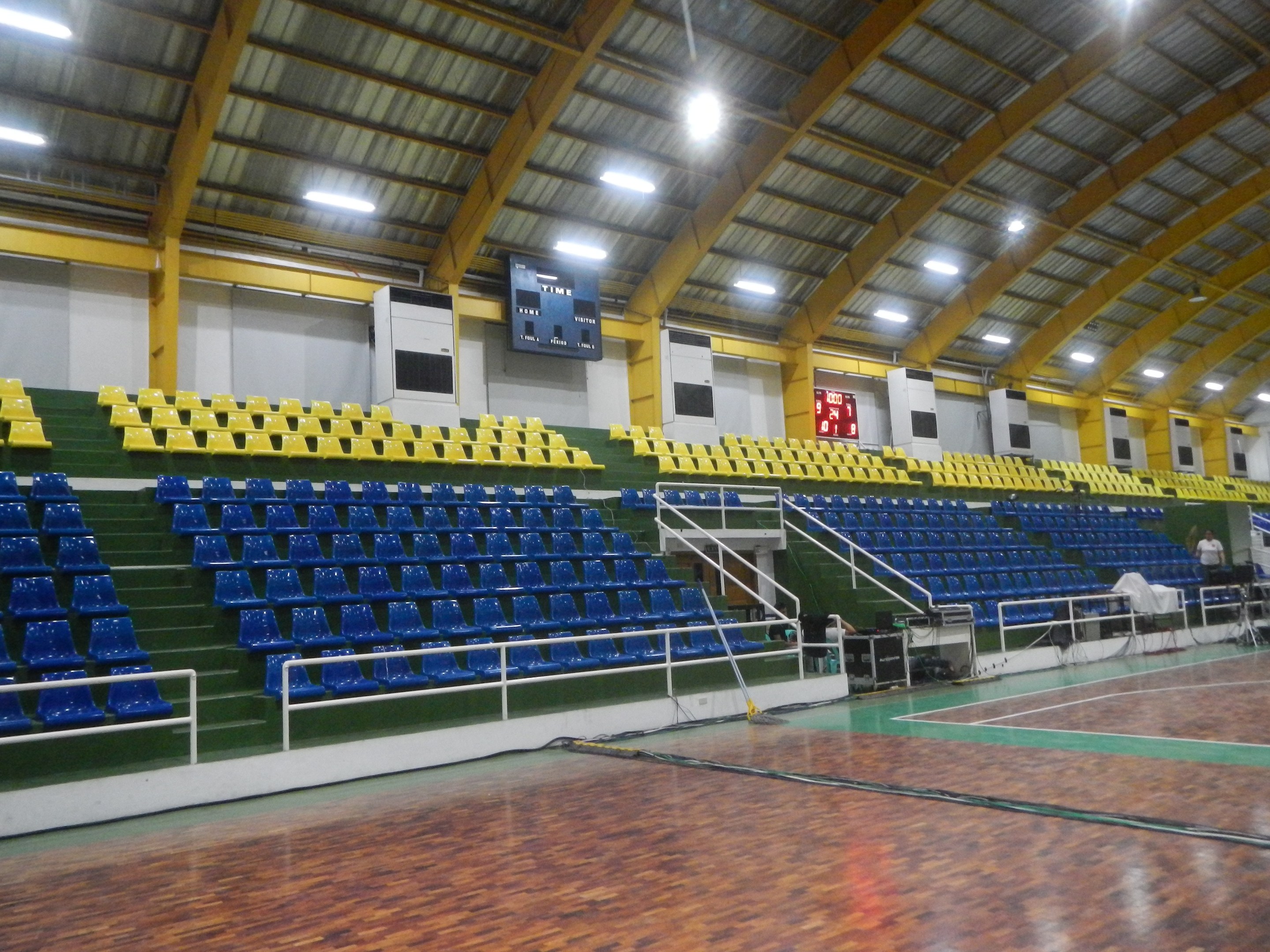 Großer Hallensportplatz mit Holzboden, blauen und gelben Sitzplätzen, Geländern, Treppen zum Sitzbereich, wandmontierten Bildschirmen, Deckenbeleuchtung und einigen Personen.