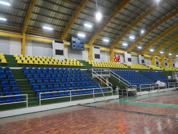 Großer Hallensportplatz mit Holzboden, blauen und gelben Sitzplätzen, Geländern, Treppen zum Sitzbereich, wandmontierten Bildschirmen, Deckenbeleuchtung und einigen Personen.