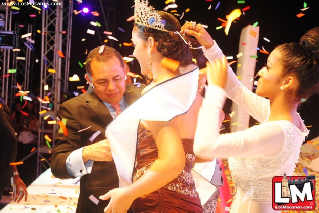 Ein Mädchen auf einer Bühne bei einer Miss-World-Show mit einer Krone, dahinter ein Mädchen, das sie anpasst, und vorne ein Mann in einem schwarzen Anzug, der die Hand schüttelt, mit Metallrahmen und dunklem Hintergrund.
