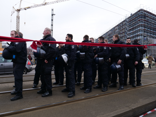 Eine Gruppe von Polizisten in schwarzen Uniformen und Helmen steht in einer Reihe auf einem Bahnsteig, mit einem roten Band davor, vor einer Kulisse aus Fahrzeugen, Passanten, Gebäuden und einem klaren Himmel.
