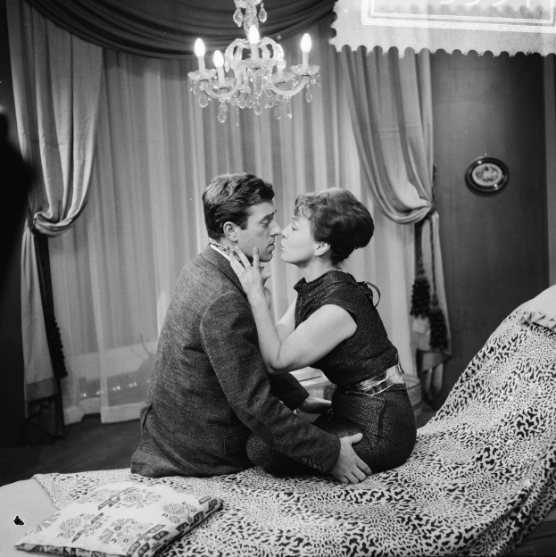 Zwei Menschen umarmen und küssen sich zärtlich auf einem Bett mit Kissen und Decke in einem sanft beleuchteten Raum mit einem Kronleuchter und Vorhängen.