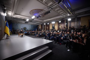 Eine grosse Gruppe von Menschen, die auf Stühlen sitzend einer Bühne mit einem zentralen Tisch, einer Fahne auf der linken Seite und Fotografen auf der rechten Seite zugewandt sind, was auf eine Pressekonferenz in der Ukraine hinweist.