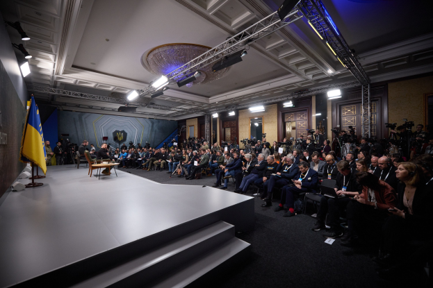 Eine grosse Gruppe von Menschen, die auf Stühlen sitzend einer Bühne mit einem zentralen Tisch, einer Fahne auf der linken Seite und Fotografen auf der rechten Seite zugewandt sind, was auf eine Pressekonferenz in der Ukraine hinweist.