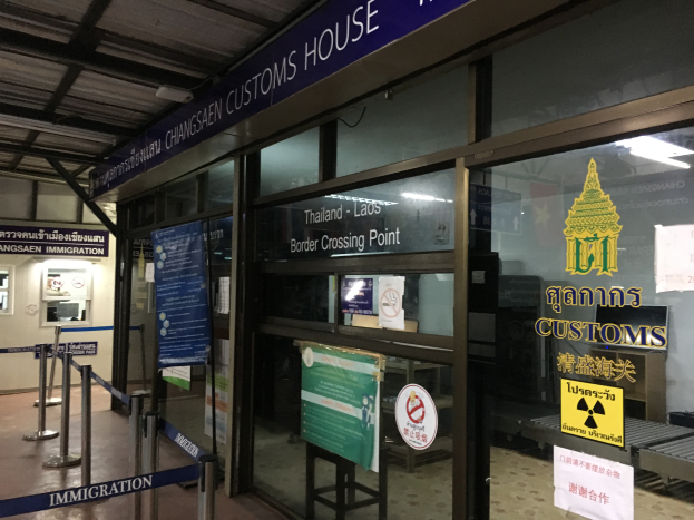 Der Eingang eines Zollhauses in Bangkok, Thailand, mit Glaswänden und -türen, Schildern mit Text und Bildern, Pfählen mit Bändern, einem Schild, einem Dach mit Deckenleuchten und einer Wand mit verschiedenen Gegenständen.