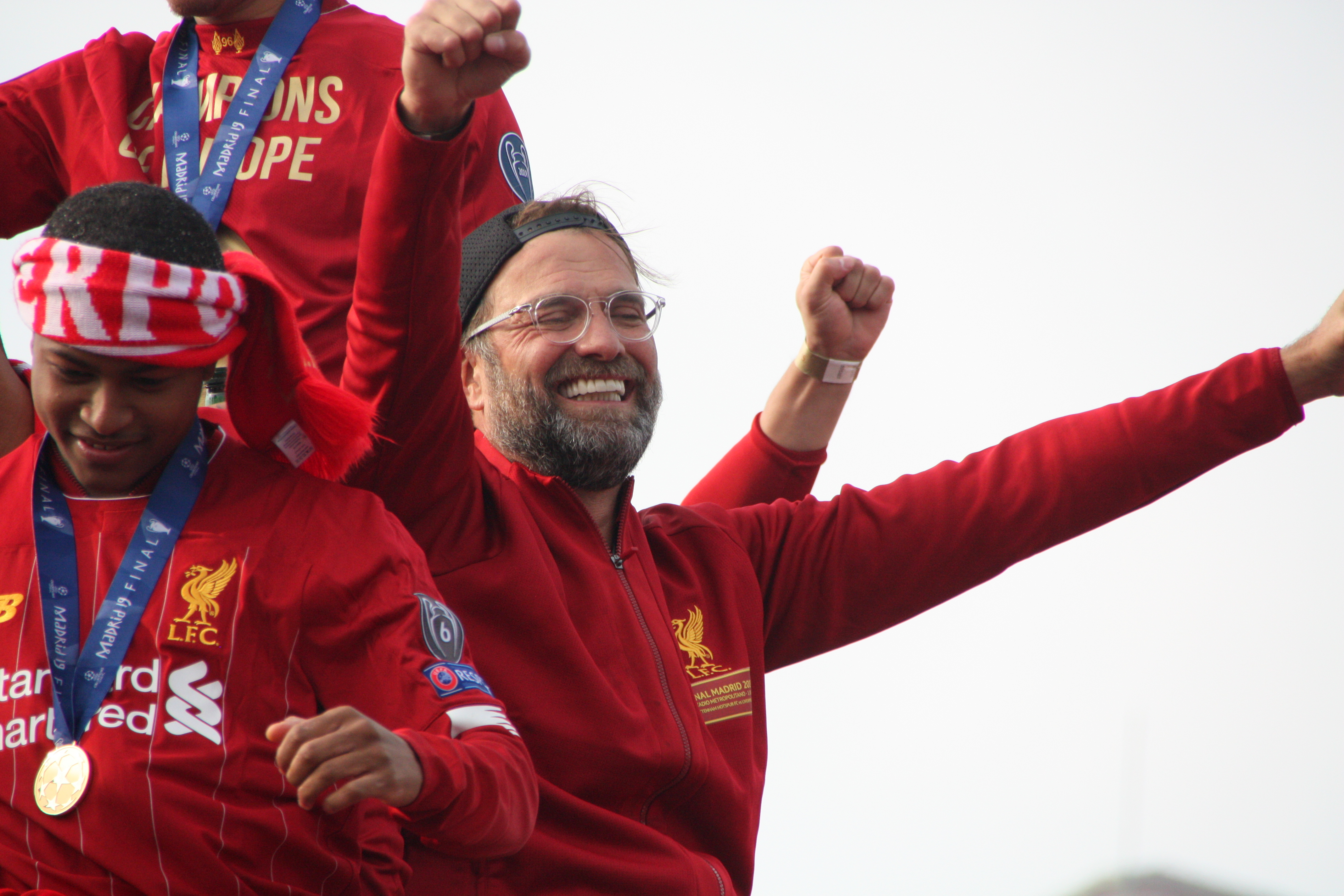 Liverpool-Trainer Jurgen Klopp feiert mit seinen Spielern den Sieg in der UEFA Champions League, während zwei mit Medaillen versehene Personen in der Nähe stehen und der Himmel im Hintergrund zu sehen ist.