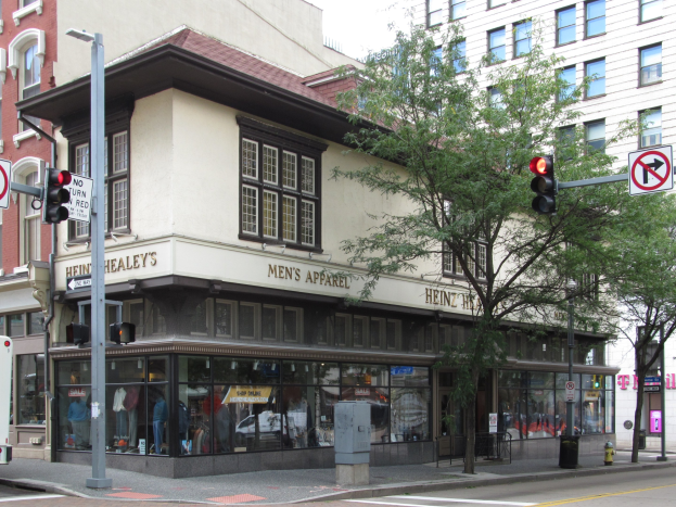 Eine Stadtstraßenecke mit einem geparkten Bus, Gebäuden, Bäumen, Laternen, Verkehrsampeln, Schildern und einem Kaufhaus mit Schaufensterpuppen.