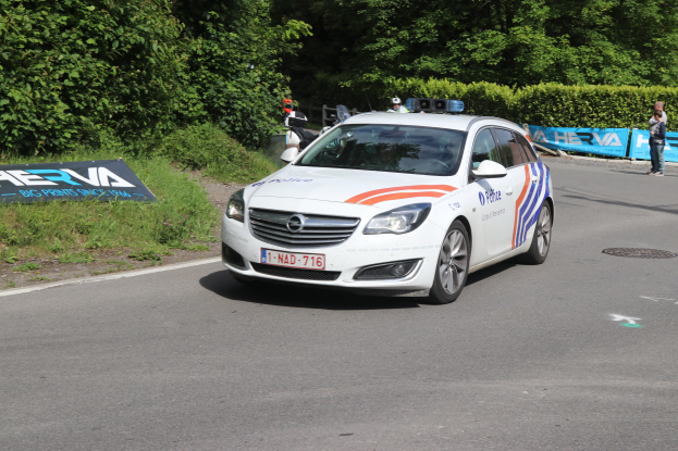 Ein Polizeiwagen fährt auf einer Straße neben einem Wald, mit Menschen auf der Seite, die eine Fahne halten, und Bäumen und Pflanzen im Hintergrund.