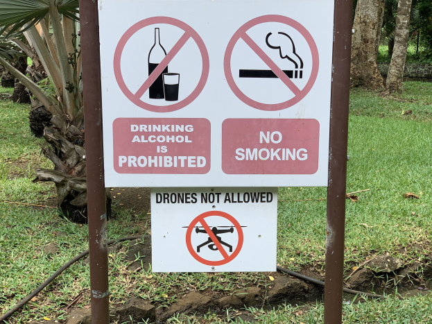 Verbotstafel am Straßenrand, die das Trinken von Alkohol und Rauchen verbietet, mit Bäumen und Gras im Hintergrund.