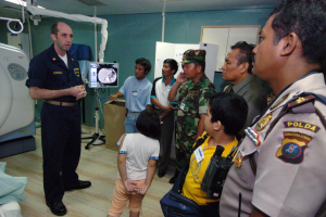 Eine Gruppe von Menschen um ein Krankenhauszimmer herumstehend, mit einem Mann in der Mitte ein Mikrofon haltend, ein Bett mit einer Decke darauf links und verschiedene medizinische Geräte im Hintergrund.