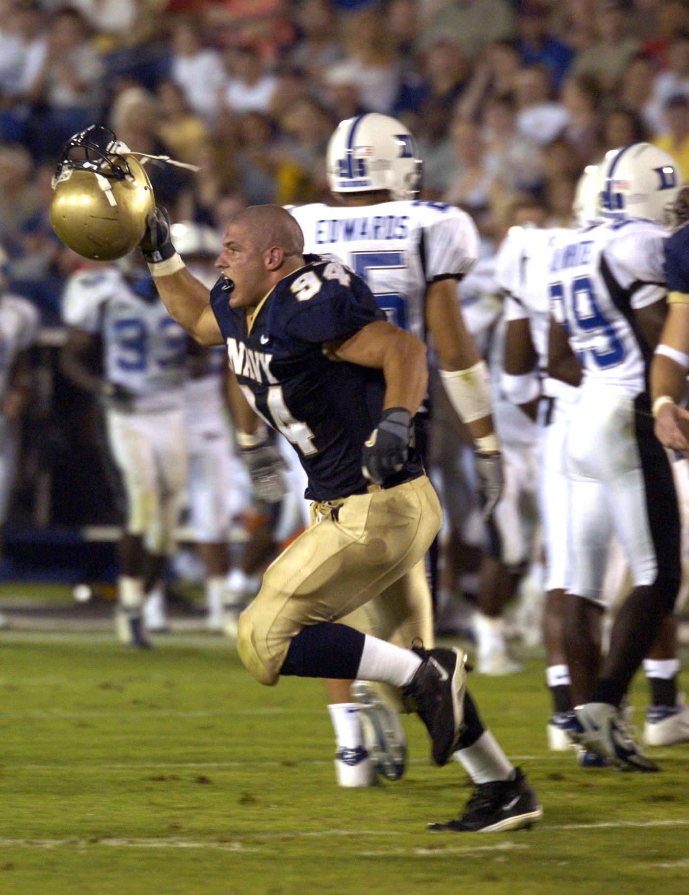 Ein Fußballspieler in dunkler Uniform rennt mit dem Ball, umgeben von Teamkollegen in ähnlicher Ausrüstung und Helmen auf dem Feld, während Zuschauer im Hintergrund auf Stühlen sitzen.