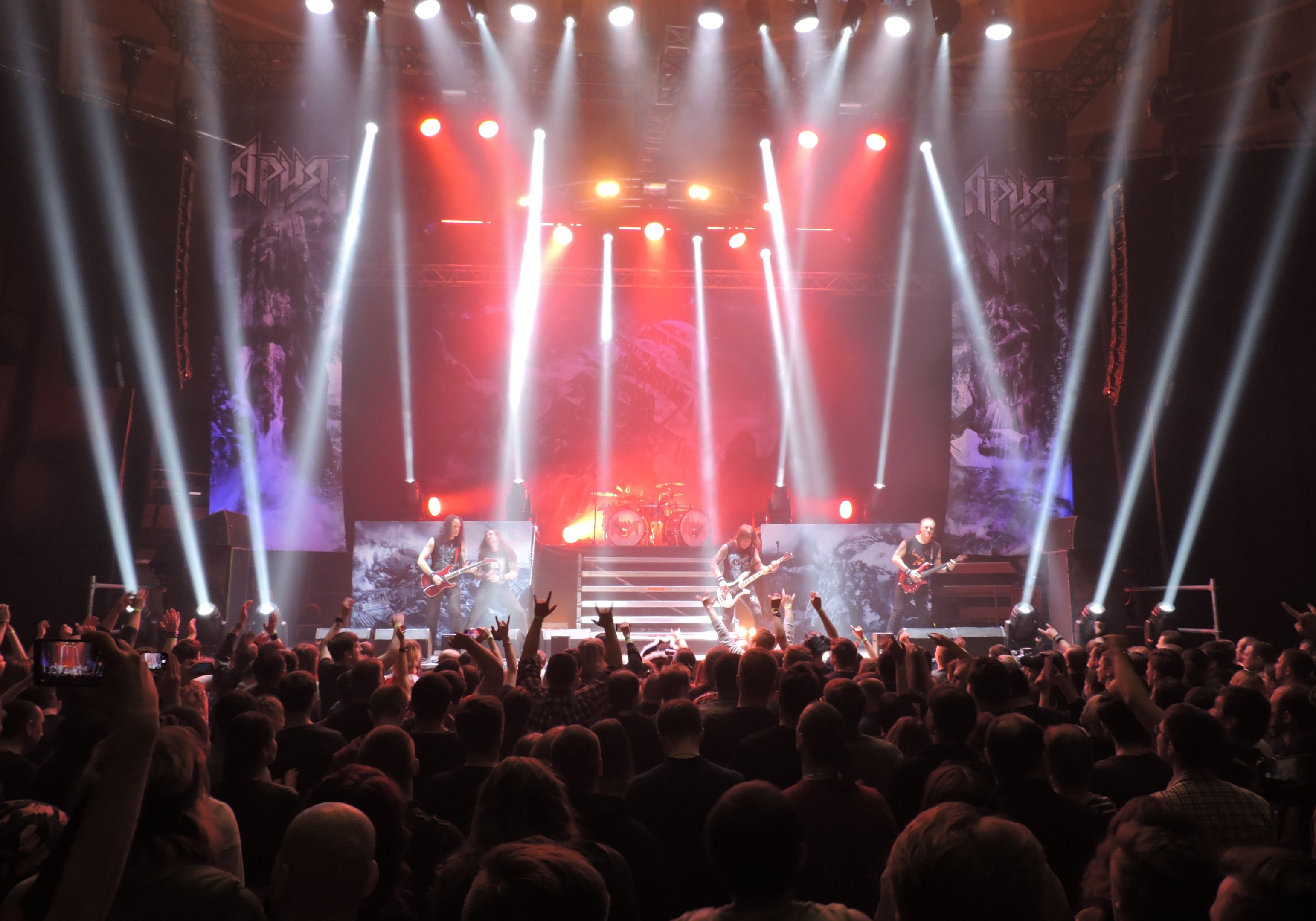 Eine Gruppe von Menschen, die auf einer hell erleuchteten Bühne spielt, einige mit Instrumenten, andere mit ihren Handys aufnehmen.