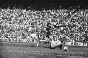 Schwarzes und weißes Foto von Fußballspielern im Einsatz auf einem Feld mit Zuschauern und einem Bundesliga 1978-79 Ajax 1-1 Hannover 96-Banner im Hintergrund.