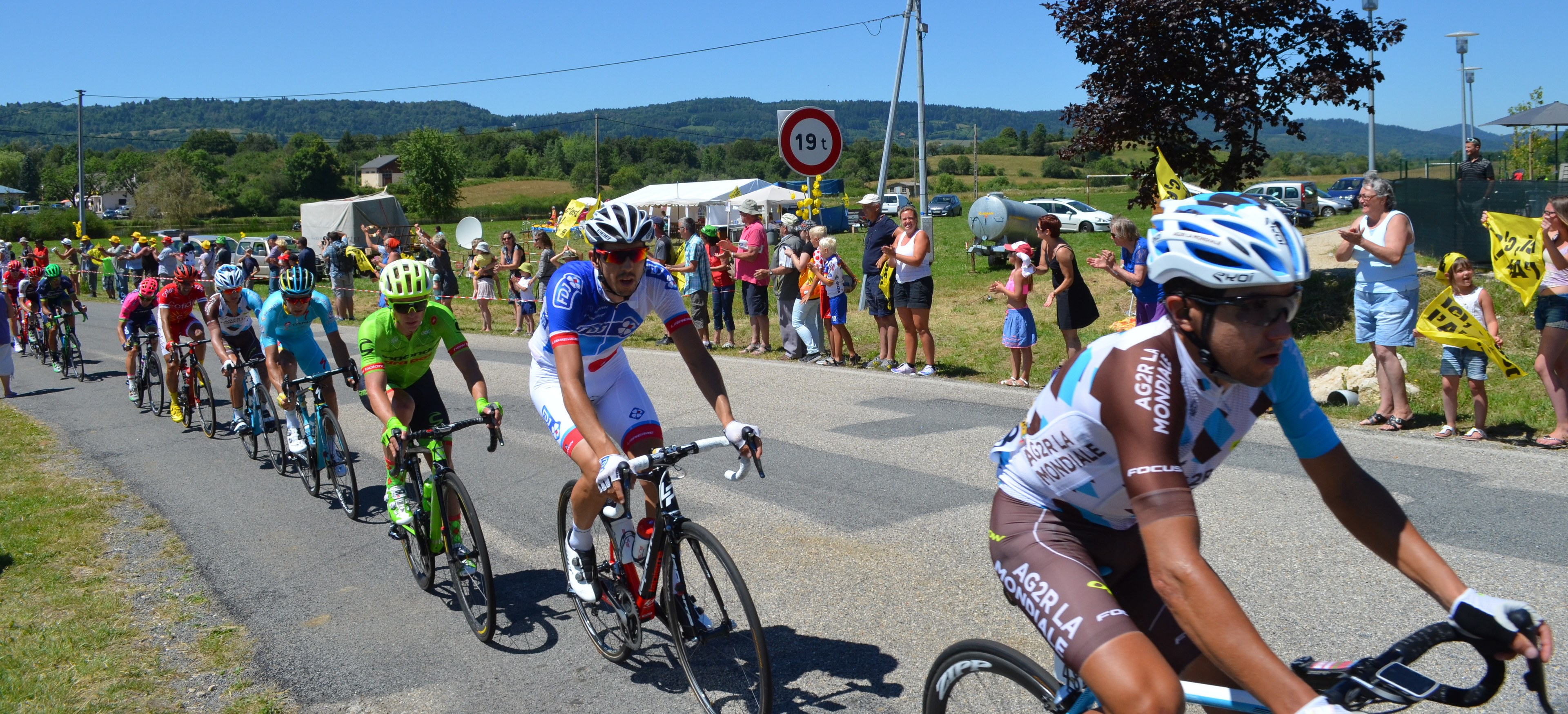Eine Gruppe von Radfahrern fährt eine Straße entlang, während Zuschauer am Straßenrand stehen, einige tragen Helme und Schutzbrillen, andere halten Fahnen, mit Bäumen, Pfählen, Schildern, Fahrzeugen, Zelten, Gebäuden, Hügeln und einem klaren blauen Himmel im Hintergrund.