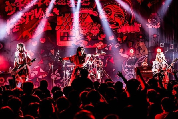 Eine Live-Aufführung der japanischen Rockband Korn auf der Bühne mit Musikern, die Gitarren spielen und singen, Mikrofonen und Ständern vorne, einer bemalten Wand im Hintergrund, Bühnenbeleuchtung und einem sichtbaren Publikum unten.