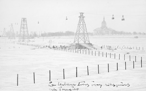 Schwarze-Weiß-Foto eines Ski-Lifts in einem verschneiten Feld mit Stützpfählen, Überseilbahn, Bäumen und einem Gebäude im Hintergrund; Text unten.