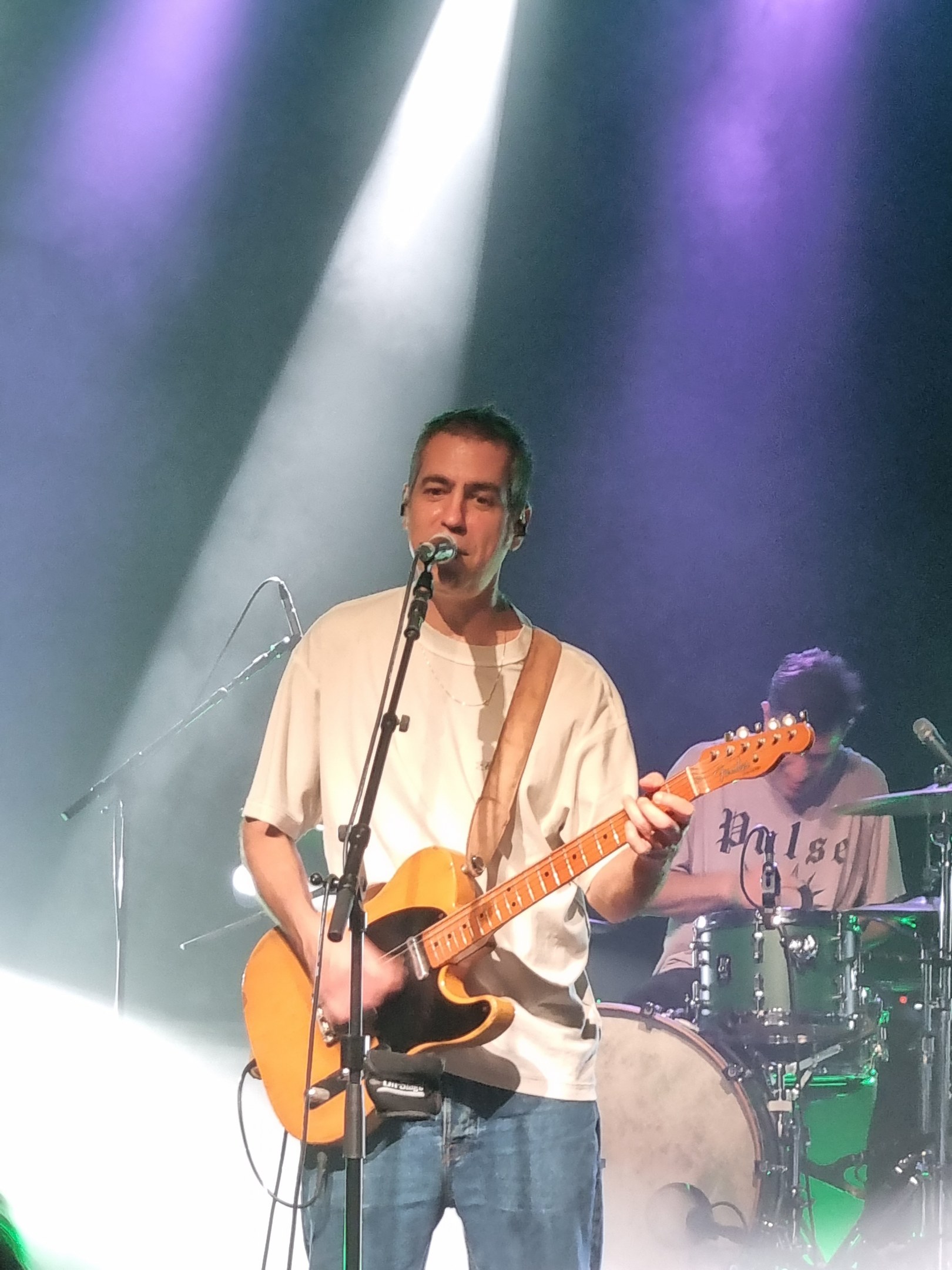 Ein Mann spielt eine E-Gitarre auf der Bühne, singt in ein Mikrofon, während ein anderer Mann Schlagzeug spielt.