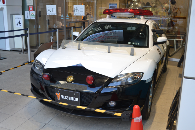 Ein Polizeiwagen steht vor einem Gebäude mit einem Bodenkegel, Absperrpfosten mit Bändern, Schildern, einer Wand, einer Tür und einem Dach mit Deckenleuchten.