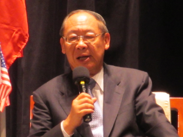 A suited man with spectacles holds a microphone while seated before a black curtain and a flag, suggesting an ambassadorial setting.