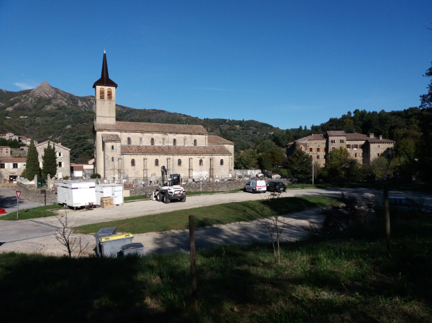 Luftaufnahme einer kleinen Stadt mit einer zentralen Kirche, umgeben von Gebäuden, Bäumen, Straßen mit Fahrzeugen und anderen städtischen Elementen, vor einem Hügel- und einem klaren blauen Himmel.