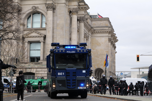 Eine Gruppe von Polizisten steht vor einem großen Gebäude mit Fenstern, Säulen und Bögen, mit Fahrzeugen auf der Straße, einer Person mit einer Kamera auf der linken Seite und einem klaren blauen Himmel mit Bäumen, Ampeln, Pfählen und Fahnen im Hintergrund.