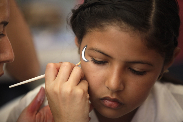 Ein Make-up-Künstler trägt Make-up auf das Gesicht eines jungen Mädchens auf, wobei beide klar gegen einen unscharfen Hintergrund zu sehen sind.